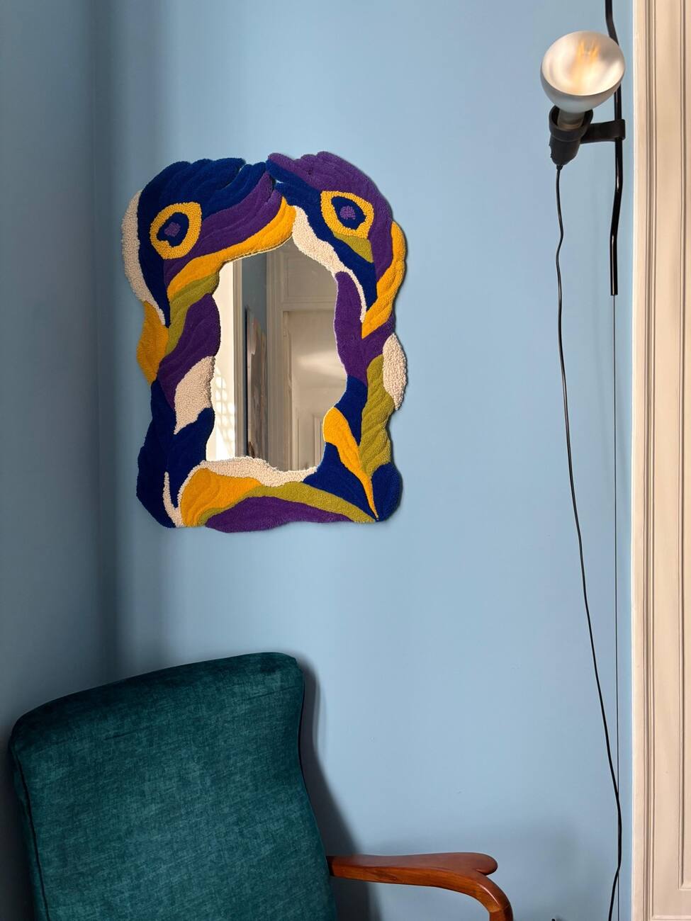 Espelho com moldura em lã azul, roxa, amarela e creme, pendurado em uma parede azul-clara ao lado de uma cadeira verde