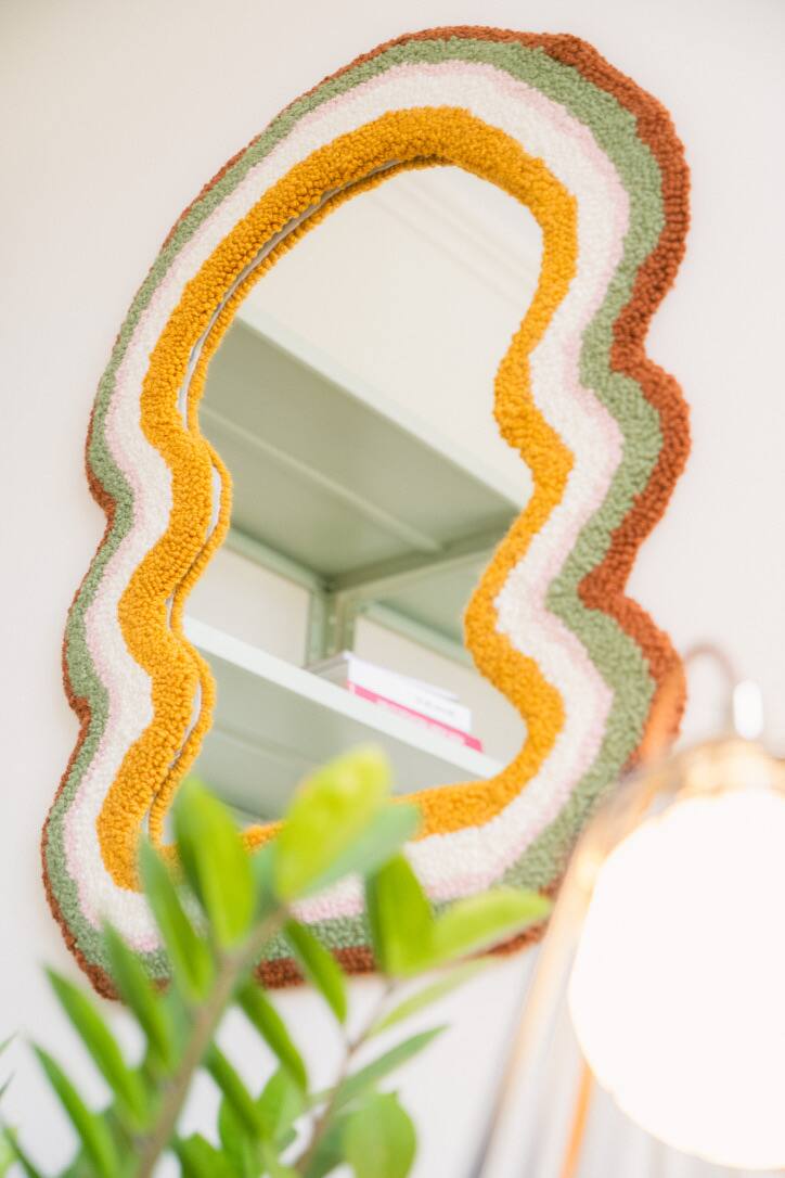 Espelho com moldura ondulada em faixas de lã amarela, cor-de-rosa, creme, verde e marrom, entre plantas