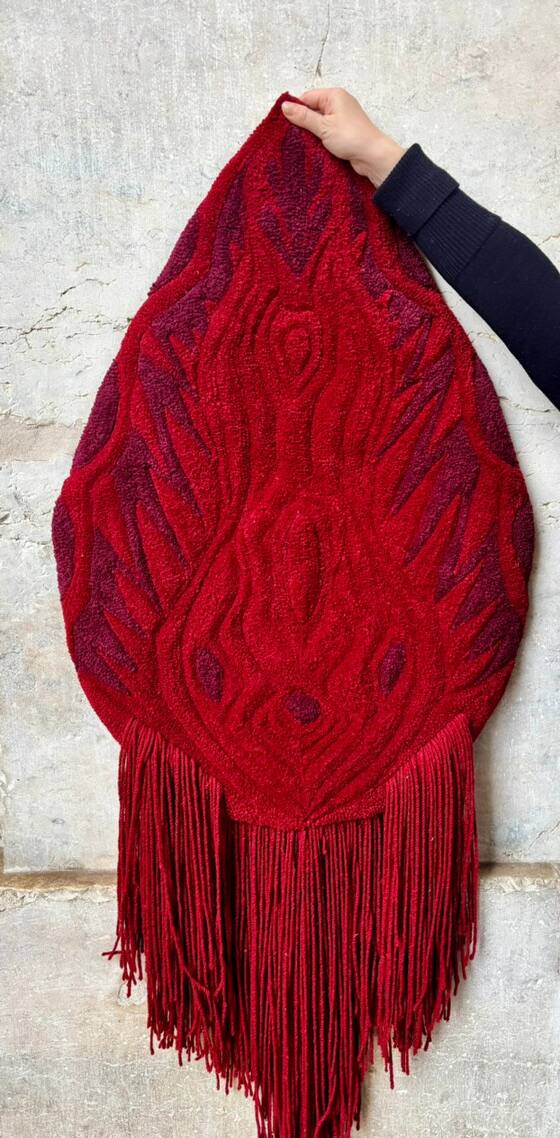 Peça em forma de gota, em lã vermelha e vermelho-escuro com franjas compridas, segurada por uma mão contra uma parede de pedra