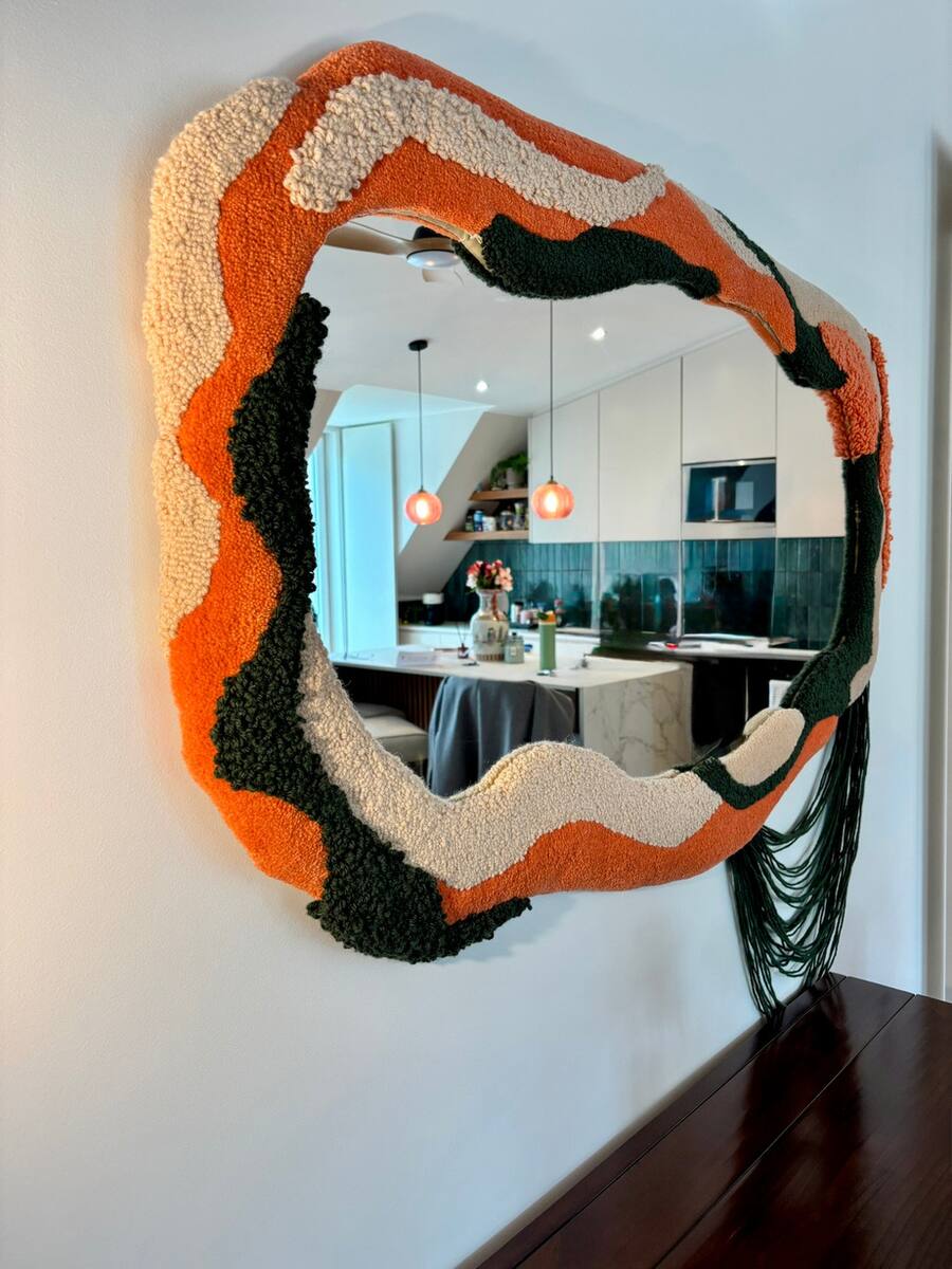 Espelho de contorno orgânico em lã laranja, creme, verde e preta, refletindo uma cozinha